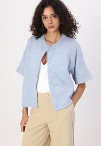Cardigan bleu clair à manches courtes en matériau doux et texturé, avec un col rond et une fermeture à boutons sur le devant.