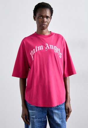 Person iført en lys pink oversized t-shirt med "Palm Angels" tekst og blå jeans med avisprint.