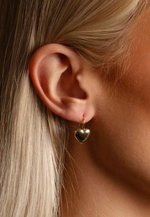 Gold heart-shaped earring with a smooth texture, hanging from a thin loop, worn on a person's ear beside straight blonde hair.