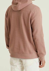 Felpa leggera rosa chiaro con superficie texturizzata, dotata di cappuccio con cordoncino, polsini a coste e tre linee di cucitura decorativa sui lati.