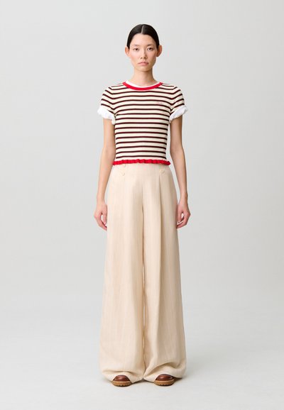 Top a maniche corte a righe con dettagli rossi e polsini bianchi, abbinato a pantaloni larghi beige con righe verticali e scarpe marroni con motivo.