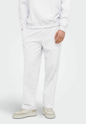 Person med hvite joggebukser, hvit genser og beige og hvite sneakers, står med en hånd i lommen.