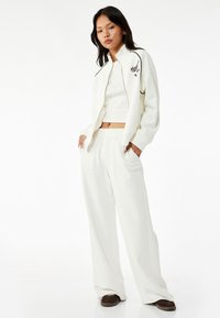 Hvid sports tracksuit med en zip-up jakke med sort kant og broderet tekst, parret med afslappede sweatpants. Blødt stof.