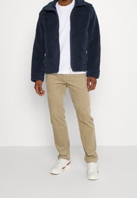 Marinblå fleecjacka med dragkedja, två sidofickor och hög krage. Bärs över en vit t-shirt och kombineras med beige kordbyxor och vita sneakers.