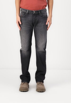 Homme portant un jean noir délavé et des chaussures à lacets marron clair, debout devant un fond clair uni.