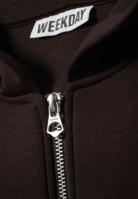 Veste zippée marron foncé avec une fermeture éclair en métal. Dispose d'un col montant et d'une étiquette portant l'inscription "WEEKDAY" sur une étiquette en tissu. Texture lisse.