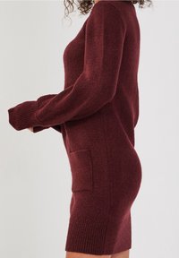 Burgundowa dzianinowa sukienka z długim rękawem, z boczną kieszenią i ściągaczami na rękawach, prezentująca teksturowany materiał i dopasowany krój.