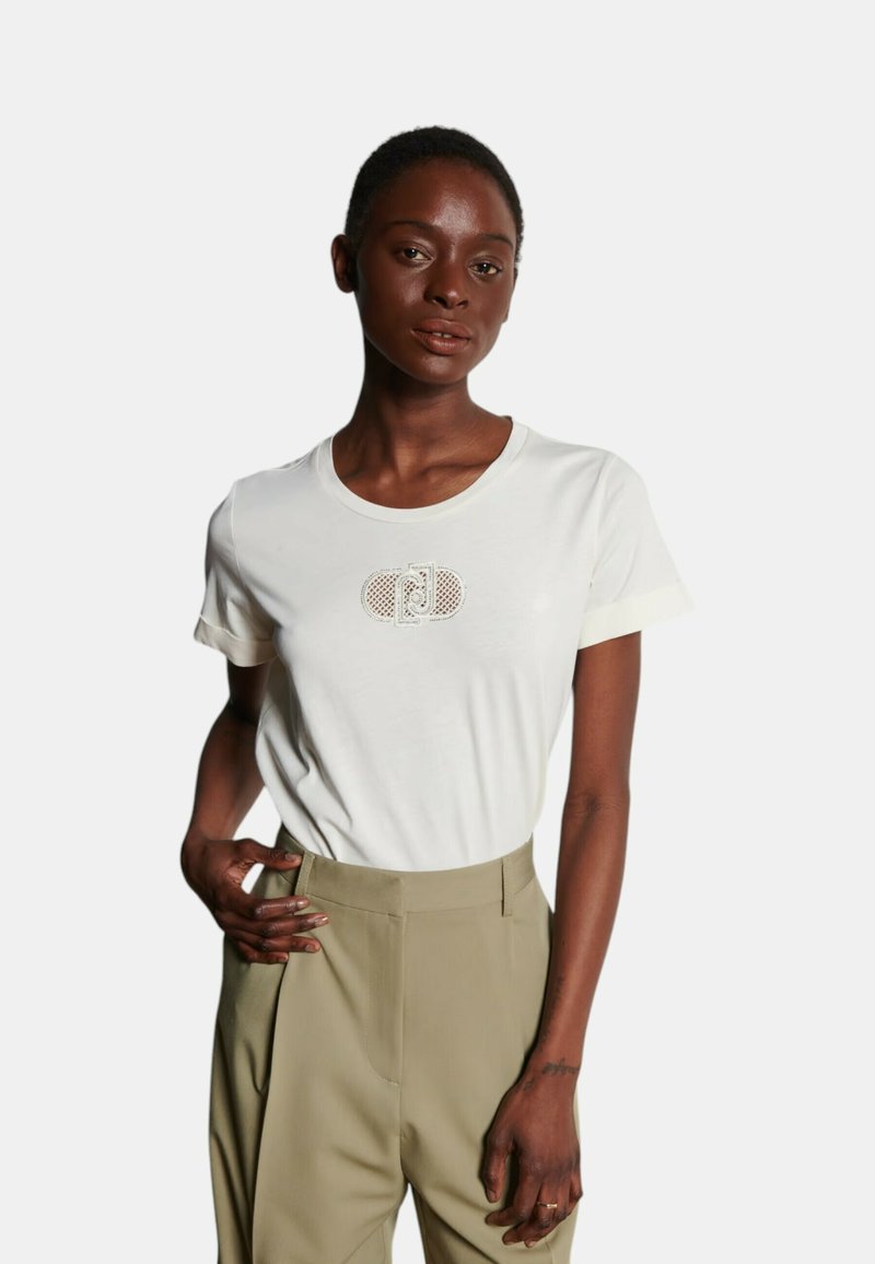 T-shirt bianca a maniche corte con un logo testurizzato al centro. Abbinata a pantaloni verde oliva, in tessuto liscio e vestibilità rilassata.
