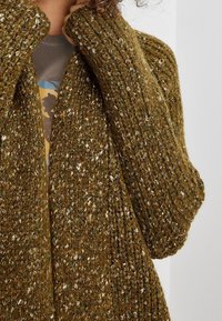 Olivgrüner Strickcardigan aus strukturiertem Garn mit metallischen Akzenten, locker geschnitten und mit weiten Ärmeln.