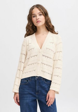 Strickjacke in Creme mit V-Ausschnitt, verziert mit dekorativen Ausschnitten und drei Knöpfen auf der Vorderseite. Lange Ärmel und gekürztes Design.