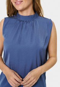 Canotta in tessuto blu morbido e liscio con design del collo arricciato e texture sottile, caratterizzata da una vestibilità rilassata e senza chiusure visibili.