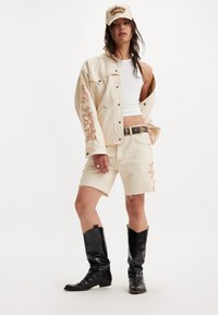 Chaqueta de mezclilla beige con diseño bordado, camiseta de tirantes blanca, pantalones cortos de mezclilla beige con dobladillo deshilachado y botas vaqueras de cuero negras. Gorra beige con texto.