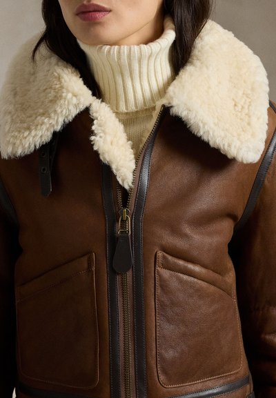 Woman wearing brown leather jacket with large cream shearling collar and cream ribbed turtleneck sweater underneath.