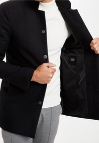 Cappotto di lana nera con chiusura a bottoni, esposto aperto per rivelare una fodera nera liscia e un'etichetta interna. Indossato sopra un collo alto bianco.