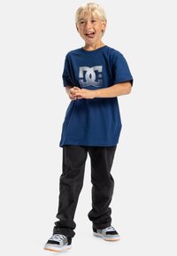 Modré bavlněné tričko s stříbrným logem DC, spárované s černými kalhotami a šedými teniskami s oranžovými akcenty a bílými tkaničkami.