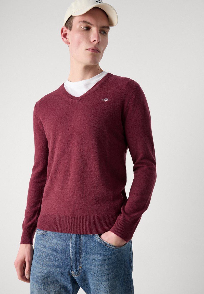 Suéter V de color marrón oscuro, hecho de un suave tejido de punto, con mangas largas y un dobladillo acanalado. Un pequeño logo está exhibido en el lado izquierdo del pecho.