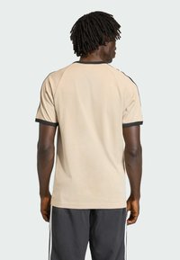 Muž s tmavou pokožkou a dredmi, oblečený v béžovom tričku s čiernym lemovaním a čiernych športových nohaviciach, stojaci chrbtom k jednoduchému pozadiu.