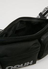 Borsa a tracolla nera con apertura con zip, parte anteriore imbottita, tracolla con logo e dettaglio del marchio. Materiale liscio con un motivo all'interno.