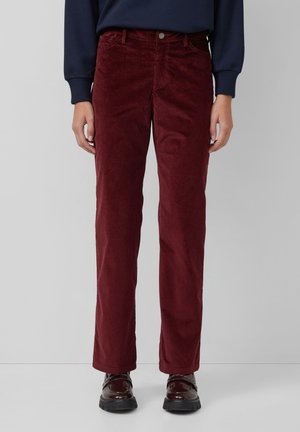 Persona che indossa pantaloni di velluto a coste color bordeaux a gamba dritta, scarpe scure lucide e una maglia blu navy a maniche lunghe su uno sfondo uniforme.