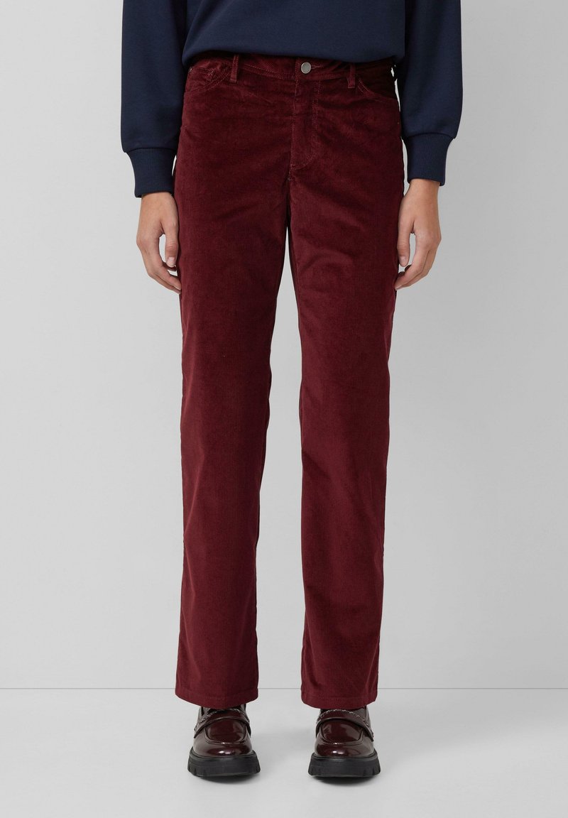 Personne portant un pantalon en velours côtelé bordeaux à jambes droites, des chaussures foncées et brillantes, et un haut bleu marine à manches longues sur un fond uni.