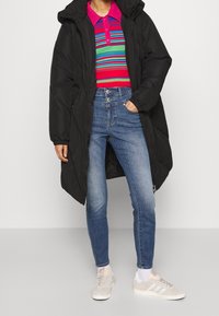 Svart puffjacka med huva, flerfärgad randig stickad polo tröja, högmidjade blå jeans och ljusgrå sneakers med vita detaljer.