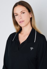 Černé polo tričko s bílým límečkem a malým vyšitým logem na levé hrudi. Má dlouhé rukávy a klasický střih.
