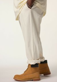 Stivali alla caviglia beige con lacci, parte superiore in pelle, collo imbottito e suola in gomma. Indossati con pantaloni crema arrotolati, mostrando texture e contrasto di colore.