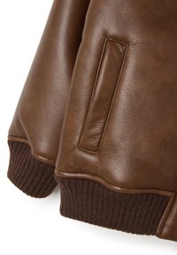 Manga de chaqueta de cuero marrón con puño de punto acanalado y detalle de bolsillo vertical en un fondo blanco.