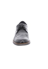 Zapato de vestir de piel negra con punta redondeada, superficie texturizada, perforaciones decorativas y una suela delgada marrón. Los cordones son negros.