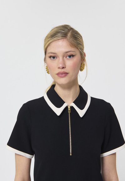 Vestido negro con mangas cortas, cuello y ribete blancos, que presenta un detalle de cremallera dorada en la parte delantera. Tejido suave, diseño clásico.