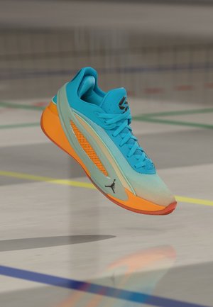 Niebiesko-pomarańczowy but sportowy w powietrzu nad halowym boiskiem sportowym z wielokolorowymi liniami, z logo Jordan po boku.