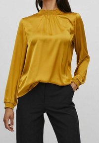 Blusa a maniche lunghe color giallo senape in satin. Presenta un collo arricciato e polsini elastici, abbinata a pantaloni neri. Texture liscia.