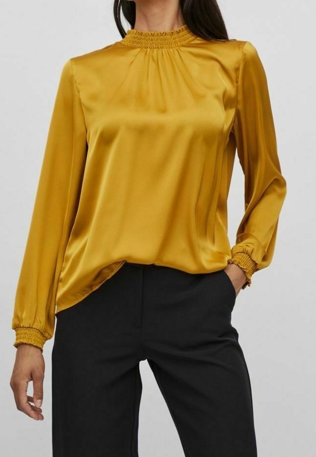 Blusa a maniche lunghe color giallo senape in satin. Presenta un collo arricciato e polsini elastici, abbinata a pantaloni neri. Texture liscia.