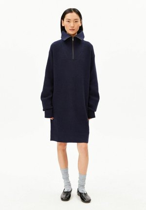 Vrouw draagt een donkerblauwe, lange mouw, gebreide jurk met een hoge ritskraag, grijze sokken en zwarte ballerina’s, staand op een eenvoudige witte achtergrond.
