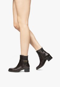 Bottines en cuir marron avec un bout rond, fermeture éclair sur le côté, et une sangle décorative avec une boucle dorée. Talon en caoutchouc, finition texturée.