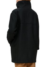 Zwarte, gestructureerde wollen jas met een opstaande kraag en naaddetails, met een rechte silhouet en verlaagde schouders, van achter gezien.