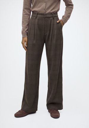 Personne portant un pantalon marron à carreaux taille haute, large, des mocassins marron et un cardigan marron clair boutonné, avec une main dans la poche.