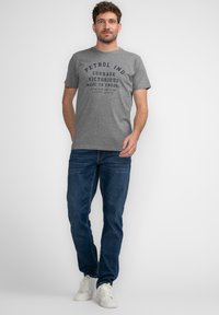 Camiseta de algodón gris con texto gráfico frontal en azul marino. Combinada con jeans azul oscuro y zapatillas blancas. Diseño sencillo y casual.