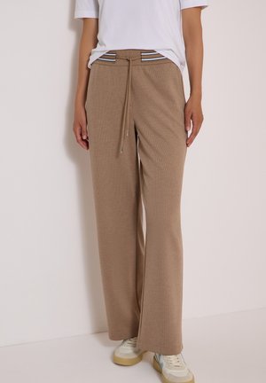 Personne portant un pantalon beige à jambes larges avec une ceinture rayée et des baskets blanches, debout devant un mur clair et uni.