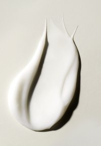 Weiße Creme oder Lotion, die in einer glatten, geschwungenen Form aufgetragen ist, mit einer glänzenden Oberfläche und leichter Textur, vor einem sanften beigen Hintergrund.