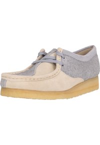 Chaussure en suède beige et gris avec un bout rond, un laçage et une semelle en caoutchouc beige. Présente des coutures visibles le long des bords.