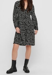 Vestido negro de manga larga con un patrón de texto manuscrito repetido, cintura ceñida y dobladillo a la altura de la rodilla. Combinado con botas negras hasta el tobillo.