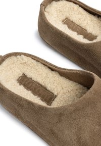 Bruine suède slippers met een zachte crème-kleurige fleece voering en een rechthoekig stoffen label binnenin. Rond gevormd en gemakkelijk aan te trekken ontwerp.