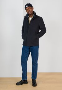 Mörk marinblå fleecejacka med hög krage, en knapp och sidofickor, i kombination med ljusblåjeans och svarta skor. Enkel design.