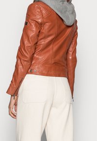 Braune Lederjacke mit eng anliegendem Schnitt, Reißverschluss an den Bündchen und einem weichen grauen Kapuzenfutter im Kragen. Getragen über hellen Hosen.