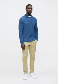 Zils teksturēts džemperis, kaki bikses un balti sporta apavi. Džempera dizainā ir apaļa apkakle un ribota aproce. Vienkāršs, mūsdienīgs dizains.