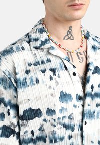 Camicia a maniche corte con una base bianca, spruzzi tinti di blu, texture plissettata e bottoni; indossata con una collana di perle multicolori.