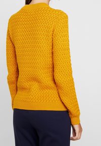 Stickad tröja i knallgult, med ett strukturerat mönster och ribbad halsringning och ärmslut. Bakåtsidan visar en figursydd design och fåll.