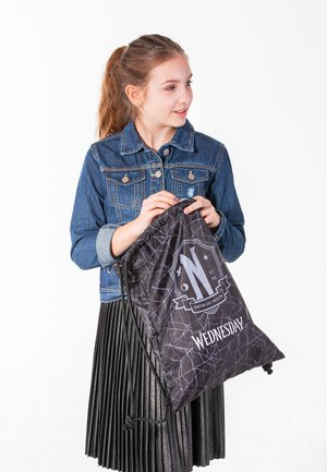 Black drawstring bag with spider web pattern and “WEDNESDAY” text. Material is shiny, durable. Girl in denim jacket holds bag, pleated skirt visible.