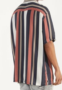 Koszula z krótkim rękawem w pionowe paski w kolorach granatowym, czerwonym, białym i beżowym, o luźnym kroju i rozcięciach po bokach. Miękka faktura materiału.